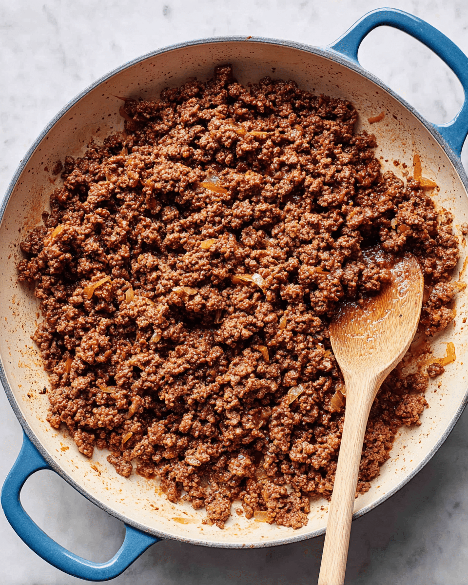 A large round white pan with blue handles holds evenly cooked ground meat, browned in color with small bits of translucent cooked onions mixed in. The ground meat is spread out in the pan, showing a crumbly texture with some darker seared spots. A wooden spoon with a light brown color lies inside the pan, slightly covered by the meat, resting on a white marbled surface. photo taken with an iphone --ar 4:5 --v 7