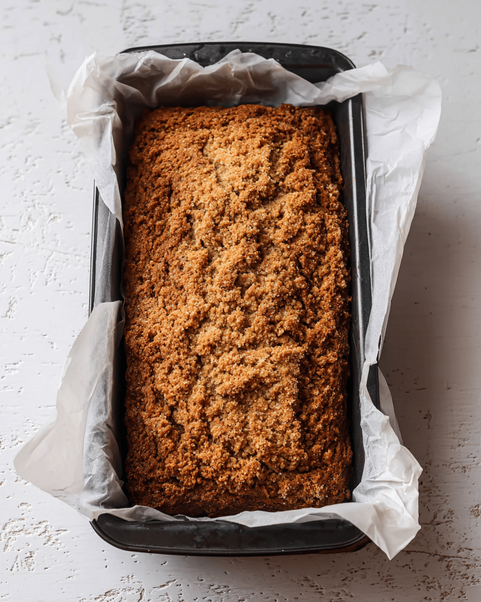 A rectangular loaf with a rough, crunchy brown top layer sits inside a black baking pan lined with white parchment paper that wrinkles and folds around the edges. The loaf's surface is uneven with a crumbly texture, showcasing small holes and cracks throughout. The baking pan rests on a white marbled textured surface, highlighting the contrast between the dark pan and the rustic-looking bread. photo taken with an iphone --ar 4:5 --v 7