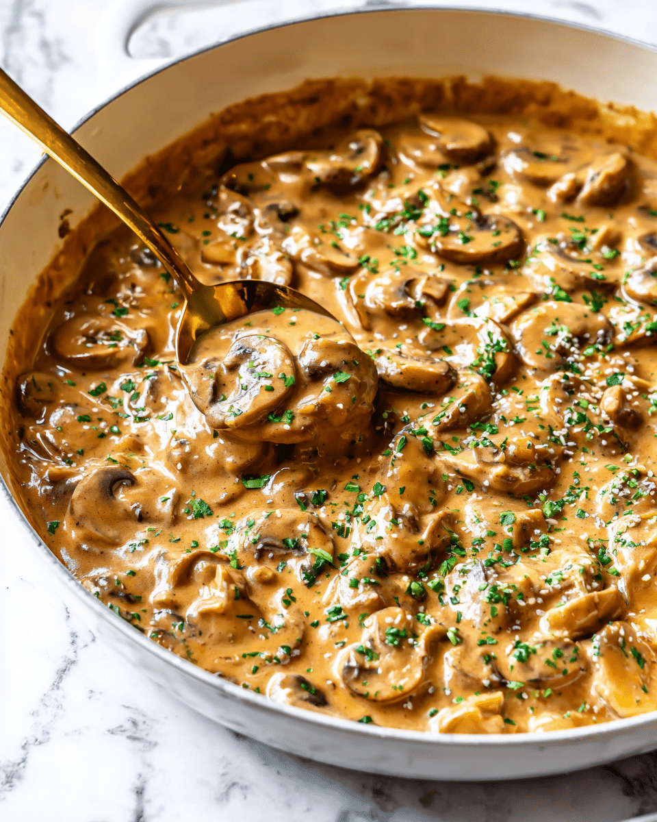 A white pan filled with creamy brown mushroom sauce, thick and smooth in texture, with many slices of soft mushrooms evenly mixed in, topped with small bits of fresh green herbs sprinkled all over. A shiny gold spoon is dipping into the sauce, lifting up a portion that shows the rich, glossy layers of mushrooms and sauce. The pan rests on a white marbled surface, creating a clean and fresh background. photo taken with an iphone --ar 4:5 --v 7