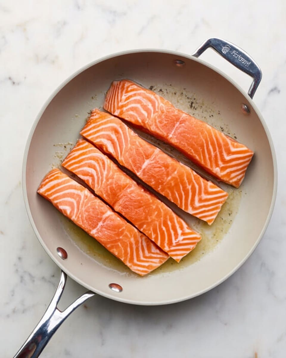 A white pan with silver handles is placed on a white marbled surface. Inside the pan, there are five pieces of raw salmon arranged side by side. The salmon pieces are pink-orange with visible lines of light fat running through them, showing a fresh, smooth texture. The pan has some light brown oil or butter shimmering around the salmon, indicating cooking has just started. photo taken with an iphone --ar 4:5 --v 7