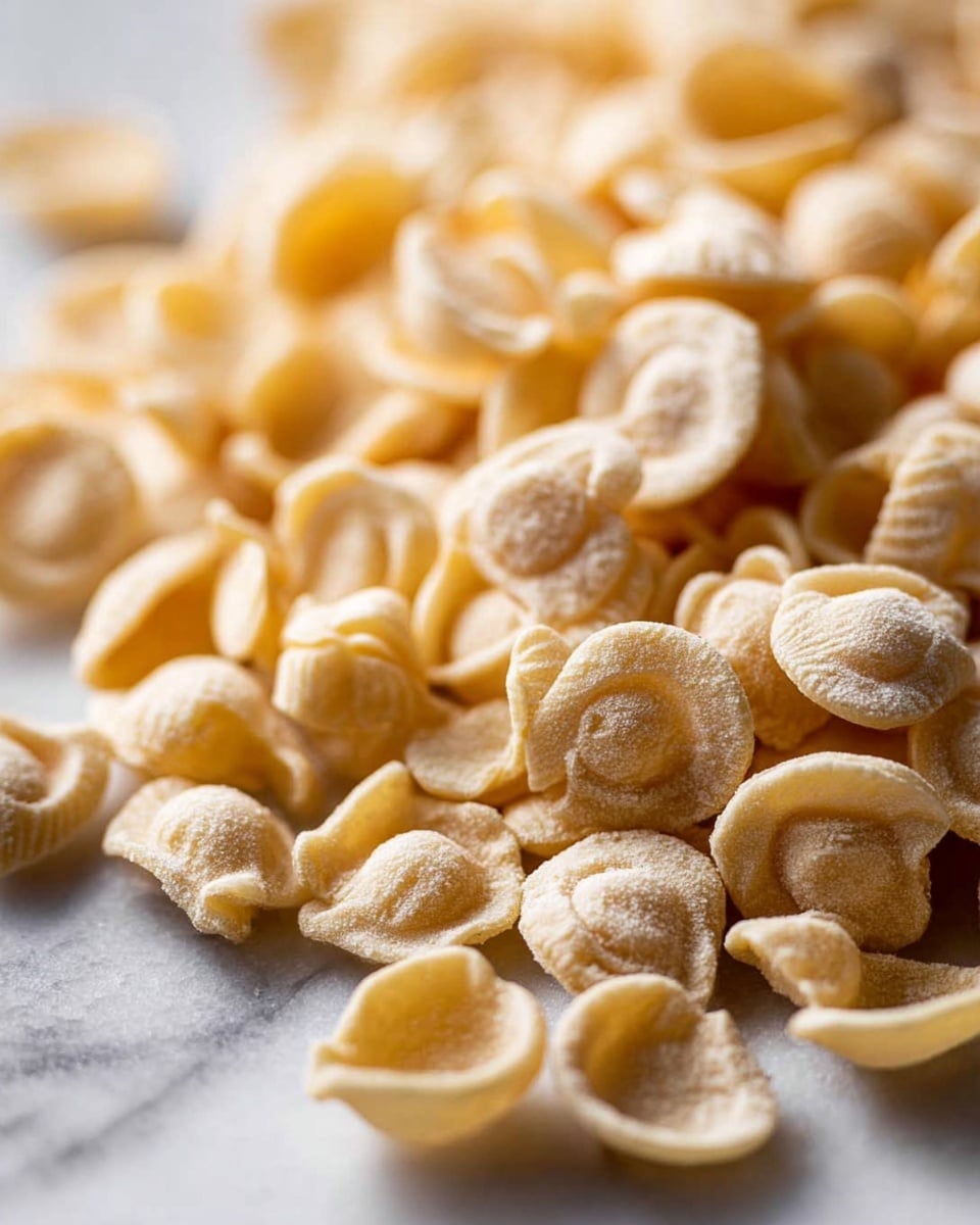 A close-up view of a pile of dry orecchiette pasta scattered on a white marbled surface, showing the small, ear-shaped pieces with a light beige color and a slightly rough texture. The pasta pieces vary in size and some show curved edges while others are more rounded, creating a natural, uneven look. The focus is sharp on the foreground pasta, while the background is softly blurred, emphasizing the shape and texture of the pasta. Photo taken with an iphone --ar 4:5 --v 7