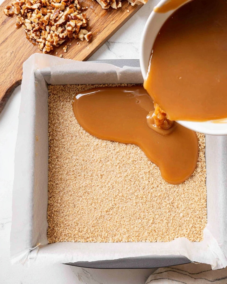 In the image, a white square baking pan lined with parchment paper holds a thick, even layer of light beige crumb crust at the bottom. From the right side, a white bowl pours a smooth, shiny caramel-colored sauce over the crust, creating a glossy puddle spreading across the surface. To the left, a wooden board with chopped nuts sits on a white marbled surface, adding a natural texture contrast. The scene is well-lit, showing a clear texture difference between the crumb base and the flowing sauce. Photo taken with an iphone --ar 4:5 --v 7