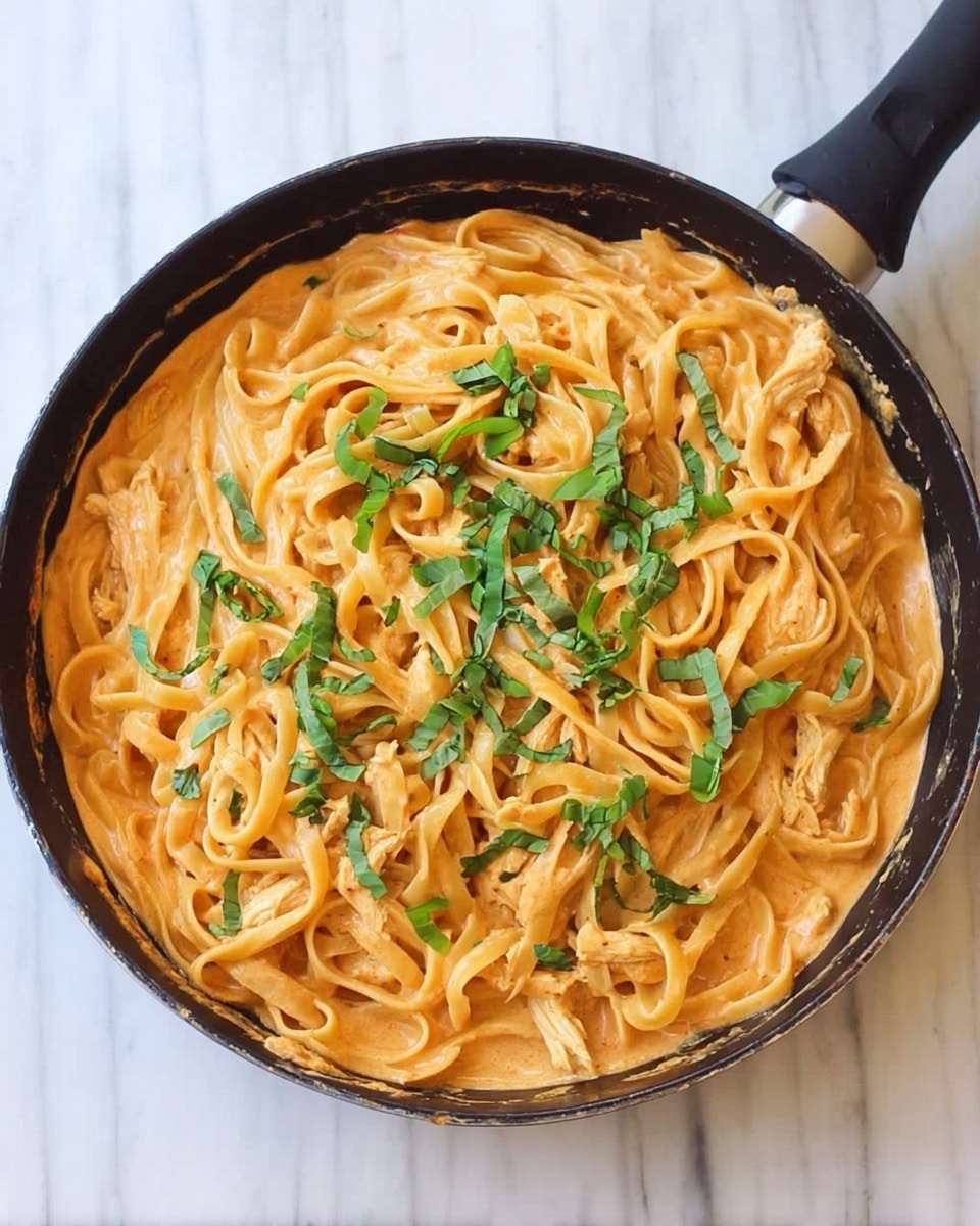 A round black pan filled with a creamy orange sauce base, topped with long, flat noodles spread evenly across the surface. Thin strips of light-colored chicken are mixed with the noodles, with fresh green basil leaves scattered on top as garnish. The pan is set on a white marbled surface. photo taken with an iphone --ar 4:5 --v 7