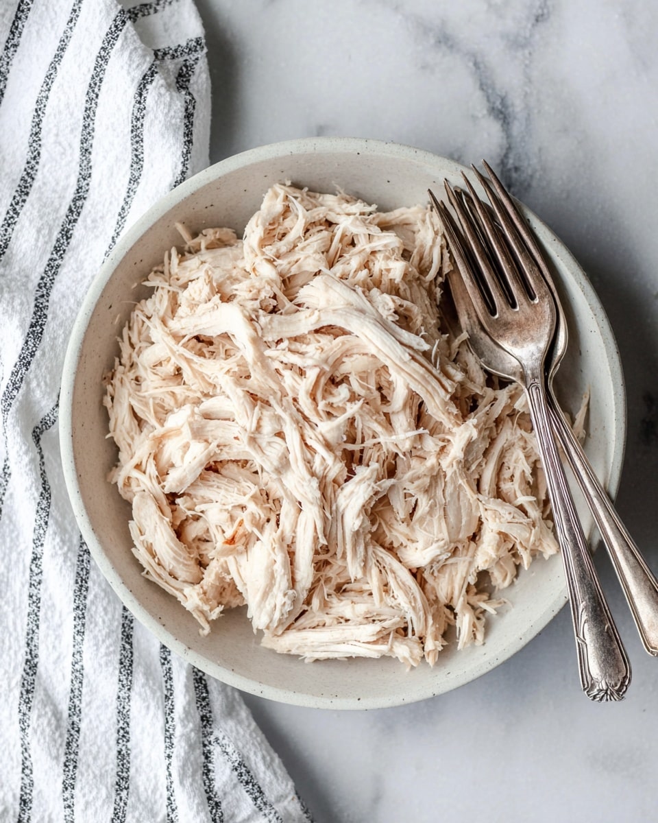 A round white bowl filled with shredded pale beige chicken pieces, showing soft, fibrous texture with some small brownish parts. Two old-fashioned silver forks rest on the right edge of the bowl. The bowl sits on a white marbled surface, with a white and black striped cloth partly visible on the bottom left corner. photo taken with an iphone --ar 4:5 --v 7