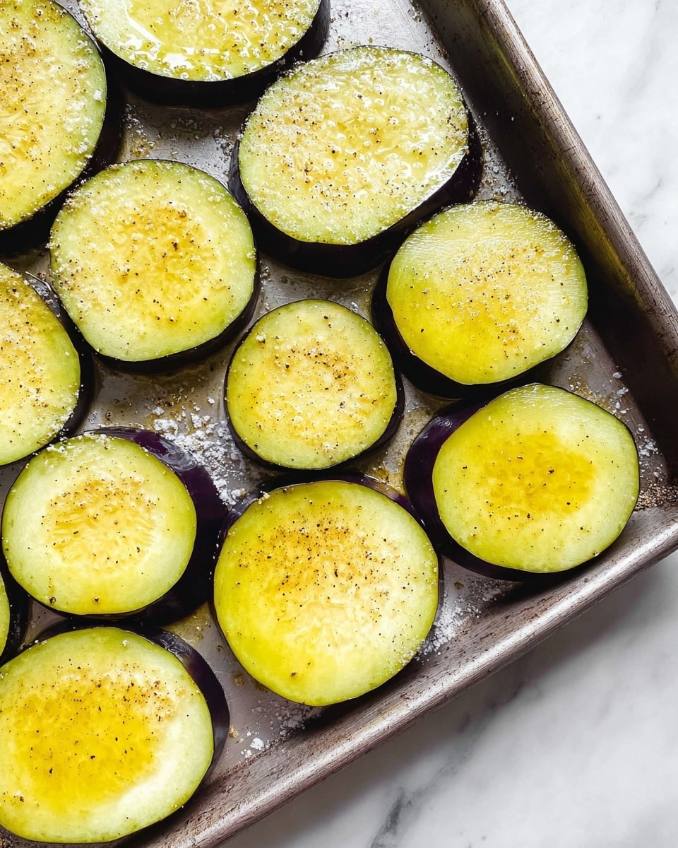 The image shows a metal baking tray filled with thick slices of eggplant. Each slice has a dark purple outer skin and a pale greenish-yellow center. The top of each slice is coated with a shiny layer of olive oil, sprinkled with small specks of black pepper and coarse salt. The tray is on a white marbled surface, and the lighting highlights the fresh texture and glossy finish of the eggplant rounds. The image captures the simplicity and freshness of the prepared eggplant ready for cooking photo taken with an iphone --ar 4:5 --v 7