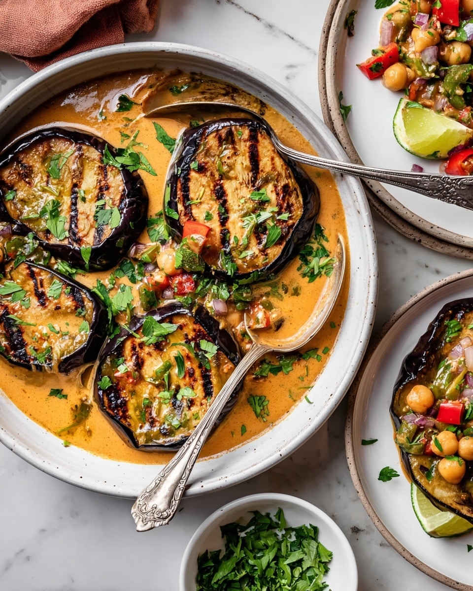 A white bowl holds four thick, grilled eggplant slices, each with dark char marks and golden brown centers, floating in a thick, creamy orange sauce with visible bits of vegetables and herbs. The eggplants are topped with fresh green parsley leaves. Two vintage silver serving spoons rest inside the bowl, partially submerged in the sauce. To the right, a white plate shows two eggplant slices with the same creamy sauce, chickpeas, diced red tomatoes, and onion pieces garnished with parsley and a lime wedge on the side. Below, a small white bowl contains finely chopped green herbs on a white marbled surface with stacked white plates nearby. Photo taken with an iphone --ar 4:5 --v 7