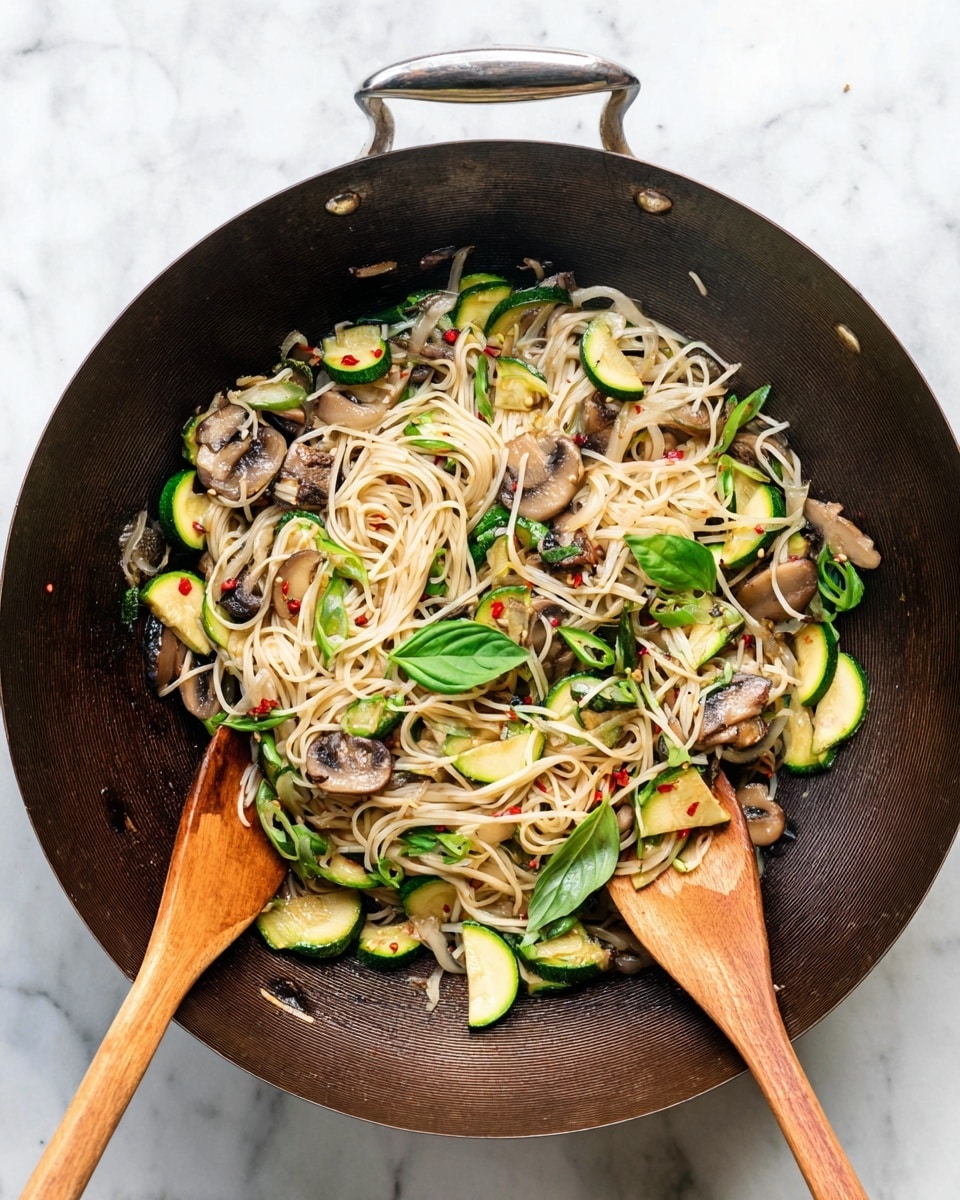 A large dark wok filled with light-colored noodles mixed with slices of green zucchini, brown mushrooms, and translucent onion strips. Bright green basil leaves and chopped green onions are scattered on top, with a few red pepper flakes adding small touches of red. Two wooden spoons rest inside the wok, one on the left and one on the right, mixing the ingredients. The wok sits on a white marbled surface. photo taken with an iphone --ar 4:5 --v 7
