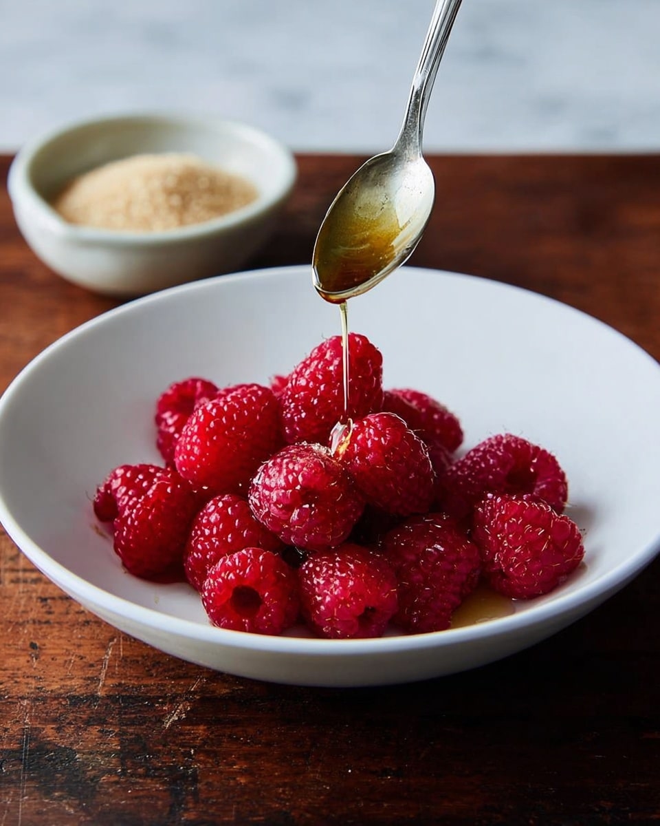 A white shallow bowl holds a small pile of bright red raspberries in the center, each berry plump and textured with tiny seeds. Above the berries, a metal spoon pours a light golden liquid, creating a shiny layer on top of the fruit. In the background, a small white bowl with light brown sugar is visible on a dark wooden surface, but the main focus is on the raspberries and the liquid being poured on them. The scene sits on a white marbled textured surface. photo taken with an iphone --ar 4:5 --v 7