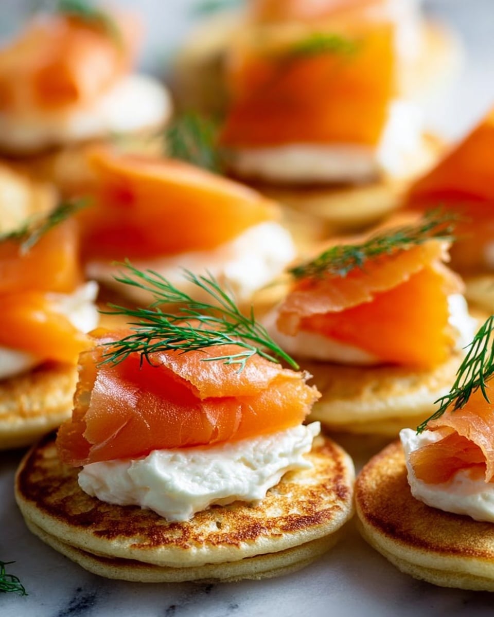 The image shows many small round white blinis spread out on a white marbled surface. Each blini has three layers: the bottom golden-brown pancake, a thick white layer of cream cheese spread with a smooth texture, and on top, an orange slice of smoked salmon shaped in a slight curl or fold. Each blini is garnished with a small green sprig of fresh dill. There is also a lemon wedge placed on the surface near some of the blinis. The colors are bright and fresh, with the orange salmon contrasting against the white cream cheese and blini. photo taken with an iphone --ar 4:5 --v 7