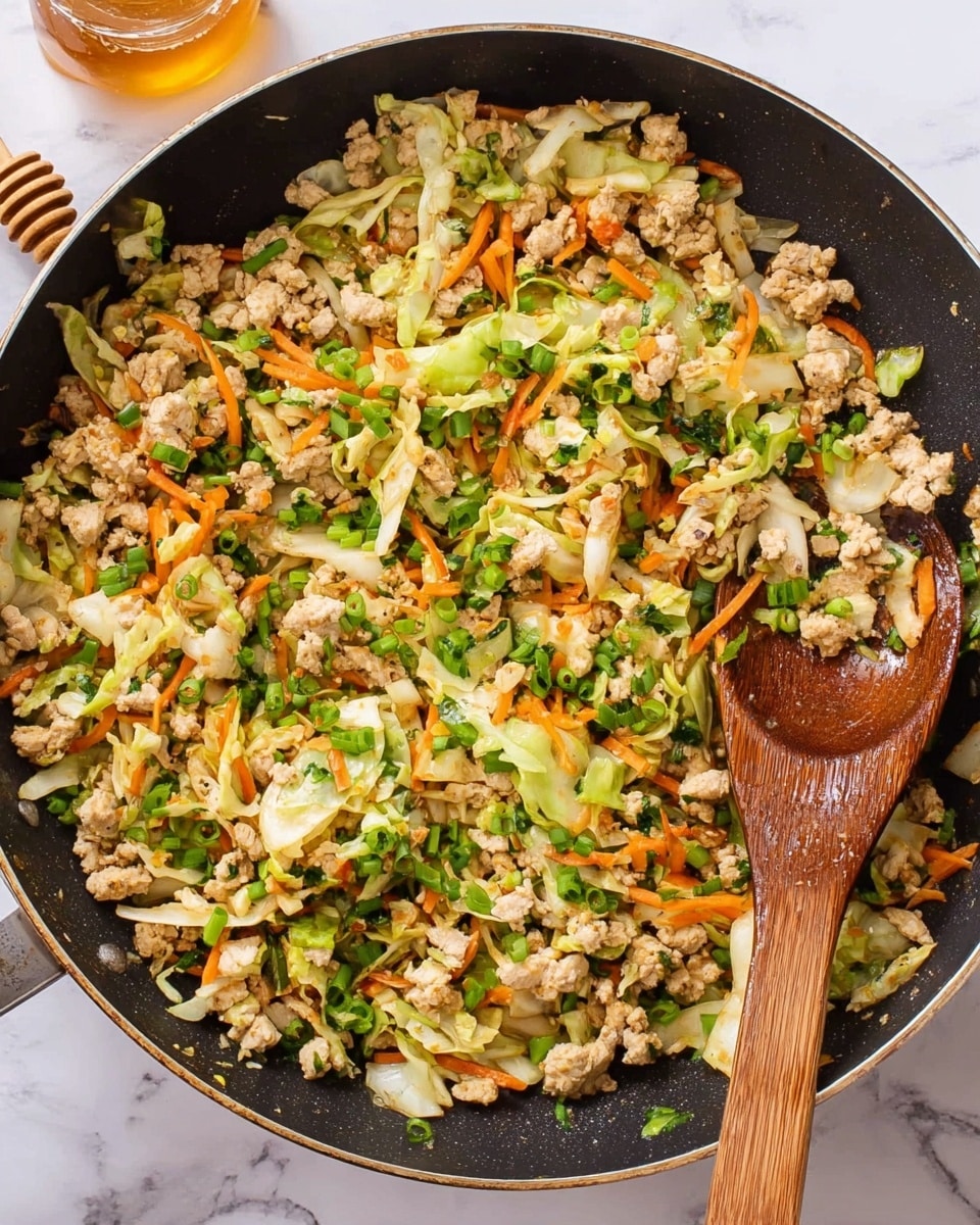 A black frying pan filled with a colorful mixture of cooked ground chicken, shredded green cabbage, thin orange carrot strips, and chopped green onions evenly spread throughout. The dish has a light texture with soft white and tan chicken pieces combined with bright green and orange vegetables, and some small bits of darker greens sprinkled on top. A wooden spoon lies on the right side of the pan, partially covered with the ingredients, showing some shine from cooking oil. The background is a white marbled surface with a honey dipper and honey jar blurred in the top left corner. Photo taken with an iphone --ar 4:5 --v 7