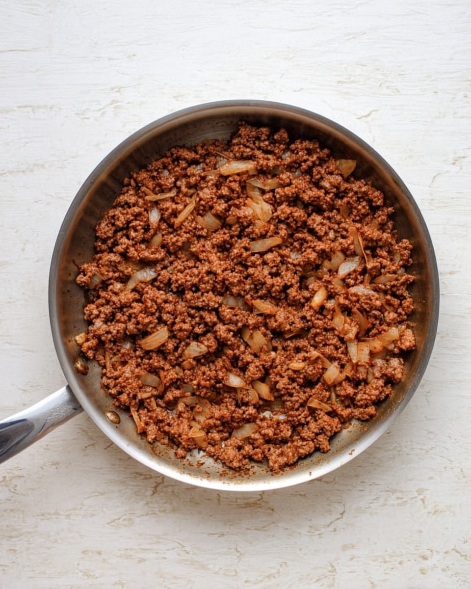 A top-down view of a round stainless steel pan filled with cooked ground meat mixed with small pieces of light brown onions. The meat is evenly browned with a slightly reddish tint from seasoning and spread across the pan surface. The pan handle extends outward and the background is a white marbled texture. photo taken with an iphone --ar 4:5 --v 7