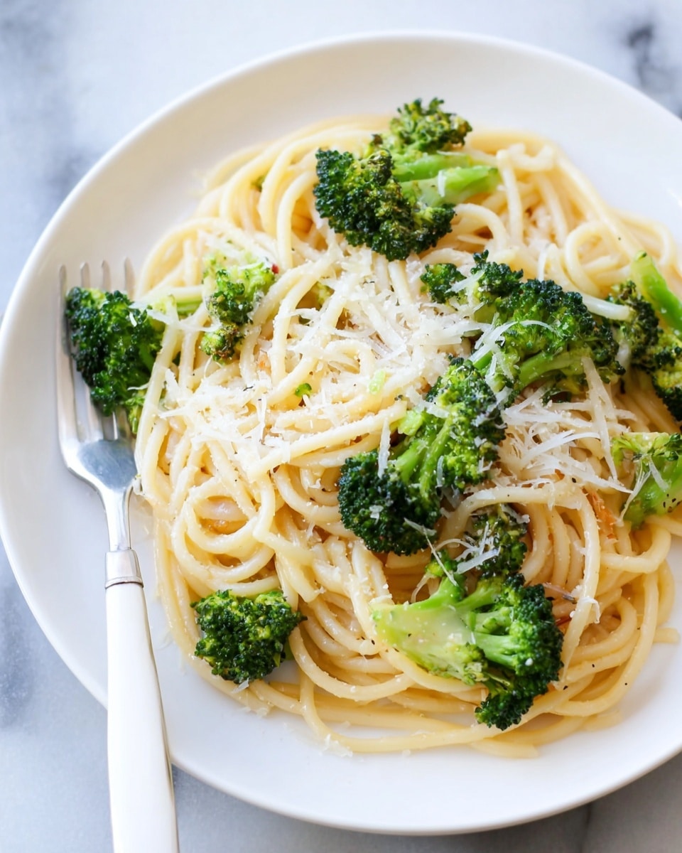 The image shows a white plate filled with one main layer of light yellow spaghetti noodles mixed with several pieces of bright green broccoli florets spread evenly throughout. The spaghetti appears soft and slightly shiny, while the broccoli looks cooked but still firm with a slightly crisp texture. On top of the noodles and broccoli, there is a light dusting of finely shredded white cheese scattered around. A silver fork with a white handle is placed on the bottom left edge of the plate, which sits on a white marbled surface. Photo taken with an iphone --ar 4:5 --v 7