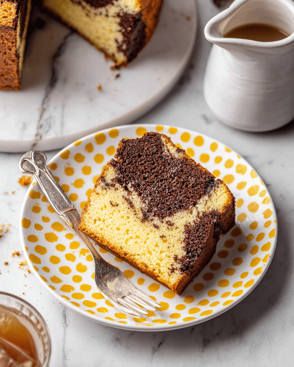 A slice of marbled cake with two distinct layers sits on a white plate with yellow polka dots around the edge; the top layer is dark brown with a slightly porous texture, and the bottom layer is light yellow with a soft crumb, blending together in the middle. A smaller cut piece lies next to the slice on the plate, with a silver fork resting nearby. The plate is placed on a white marbled surface, with part of a glass of iced coffee and a white ceramic pitcher visible at the top edges of the image. photo taken with an iphone --ar 4:5 --v 7