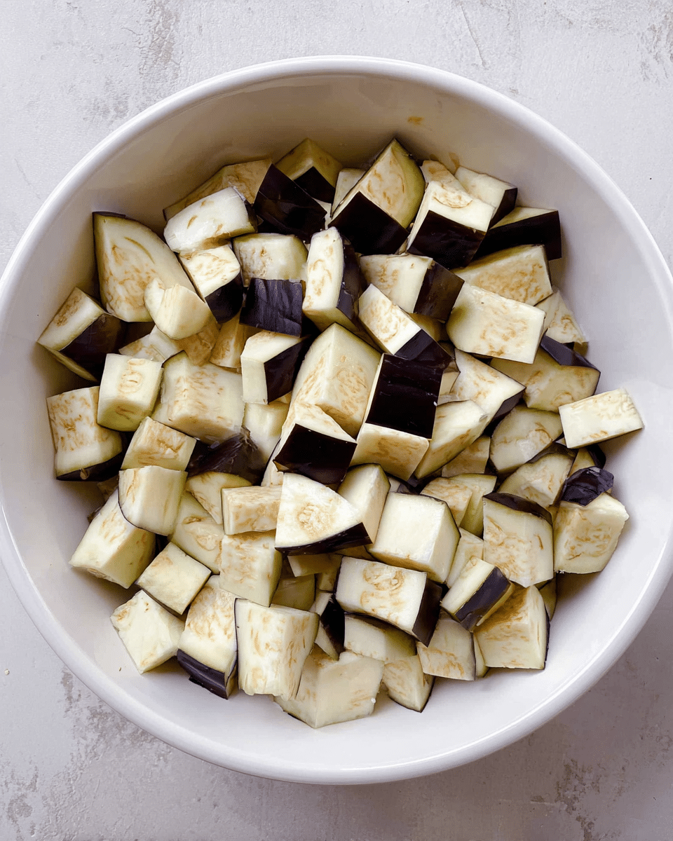 A white bowl filled with many pieces of chopped eggplant, showing creamy white flesh with small brown seeds inside and dark purple skin on some edges, all piled evenly inside the bowl; the bowl sits on a white marbled surface photo taken with an iphone --ar 4:5 --v 7