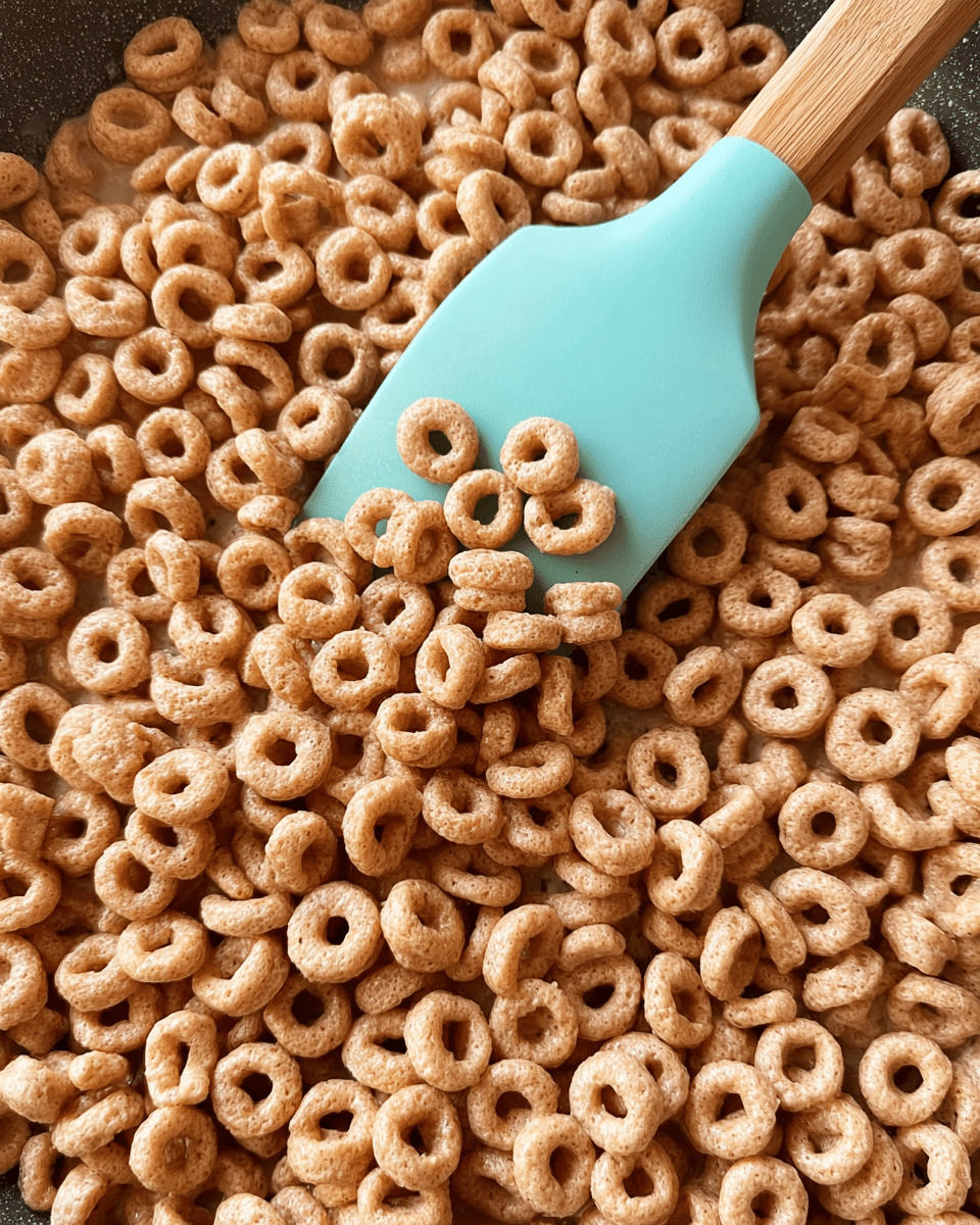The image shows a pan filled with a large amount of light brown ring-shaped cereal pieces. A light blue spatula with a wooden handle is lifting some cereal in the center, showing the texture of the small crunchy circles that look slightly puffy and have a matte surface. The background is a white marbled texture. photo taken with an iphone --ar 4:5 --v 7