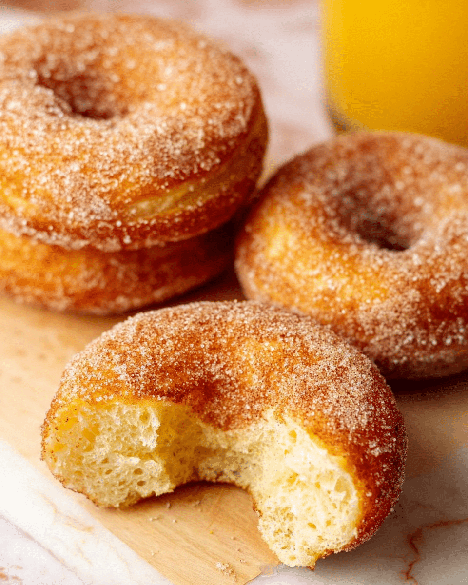 The image shows three round cinnamon sugar-coated donuts placed on a light wooden surface with a white marbled texture underneath. One donut is in the front and center, slightly cut open to reveal the soft, fluffy, and light yellow inside with a spongy texture. The other two donuts are positioned behind it, one fully visible and the other partially, all covered evenly with granulated sugar and cinnamon. A clear glass of orange juice is visible to the right of the donuts, adding a refreshing color contrast to the warm tones of the donuts. The photo is taken very close to the donuts, highlighting their texture and sugar coating. Photo taken with an iphone --ar 4:5 --v 7