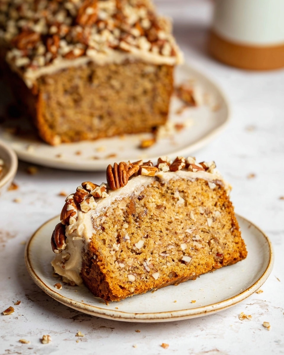 The image shows a thick slice of nut bread on a white plate with a subtle rustic edge. The bread has a light brown color with visible chopped nuts inside and throughout the slice, giving it texture. The bottom edge of the bread is covered with a layer of creamy frosting, light beige in color, which is decorated with chopped pecans pressed into it. Behind the slice, the remaining loaf is visible on another white plate, showing the same nut-studded interior and a crumbly top sprinkled with chopped nuts. The whole scene is set on a white marbled surface with soft natural lighting that highlights the warm tones and textures of the bread and nuts. photo taken with an iphone --ar 4:5 --v 7