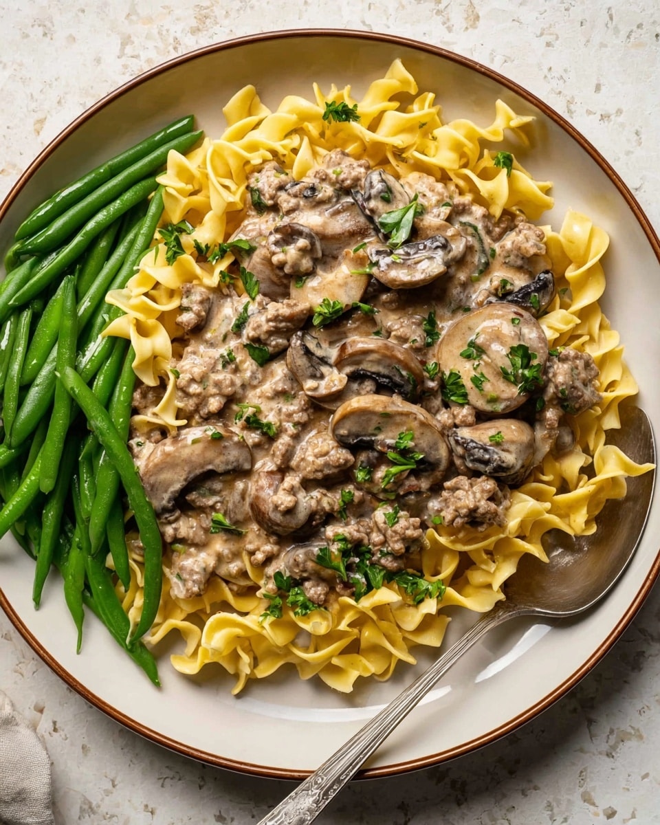 A white round plate with a brown rim holds a creamy beef stroganoff dish. The first layer is a bed of yellow egg noodles with a soft texture and twisted shapes. On top, there are chunks of browned ground beef mixed with creamy, light brown mushroom sauce that coats the noodles. Large slices of cooked mushrooms, dark beige with soft edges, are spread throughout. Fresh green parsley leaves are sprinkled over the dish, adding a touch of color. On the left side of the plate, there is a small pile of fresh green beans, bright and slightly shiny. A silver spoon rests on the right side of the plate, partially under the pasta. The plate is placed on a white marbled surface photo taken with an iphone --ar 4:5 --v 7