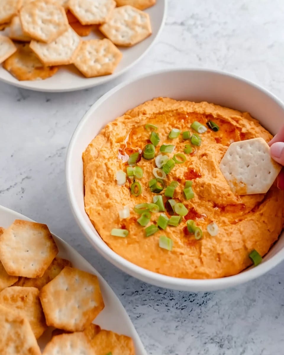 The image shows a white bowl filled with an orange creamy dip, which appears smooth with some small chunks. The dip is topped with small green sliced scallions scattered on the surface. A woman's hand is holding a white hexagon-shaped cracker, dipping it into the orange dip. Around the main bowl, there are more white plates with several hexagon-shaped crackers, placed on a white marbled surface. photo taken with an iphone --ar 4:5 --v 7