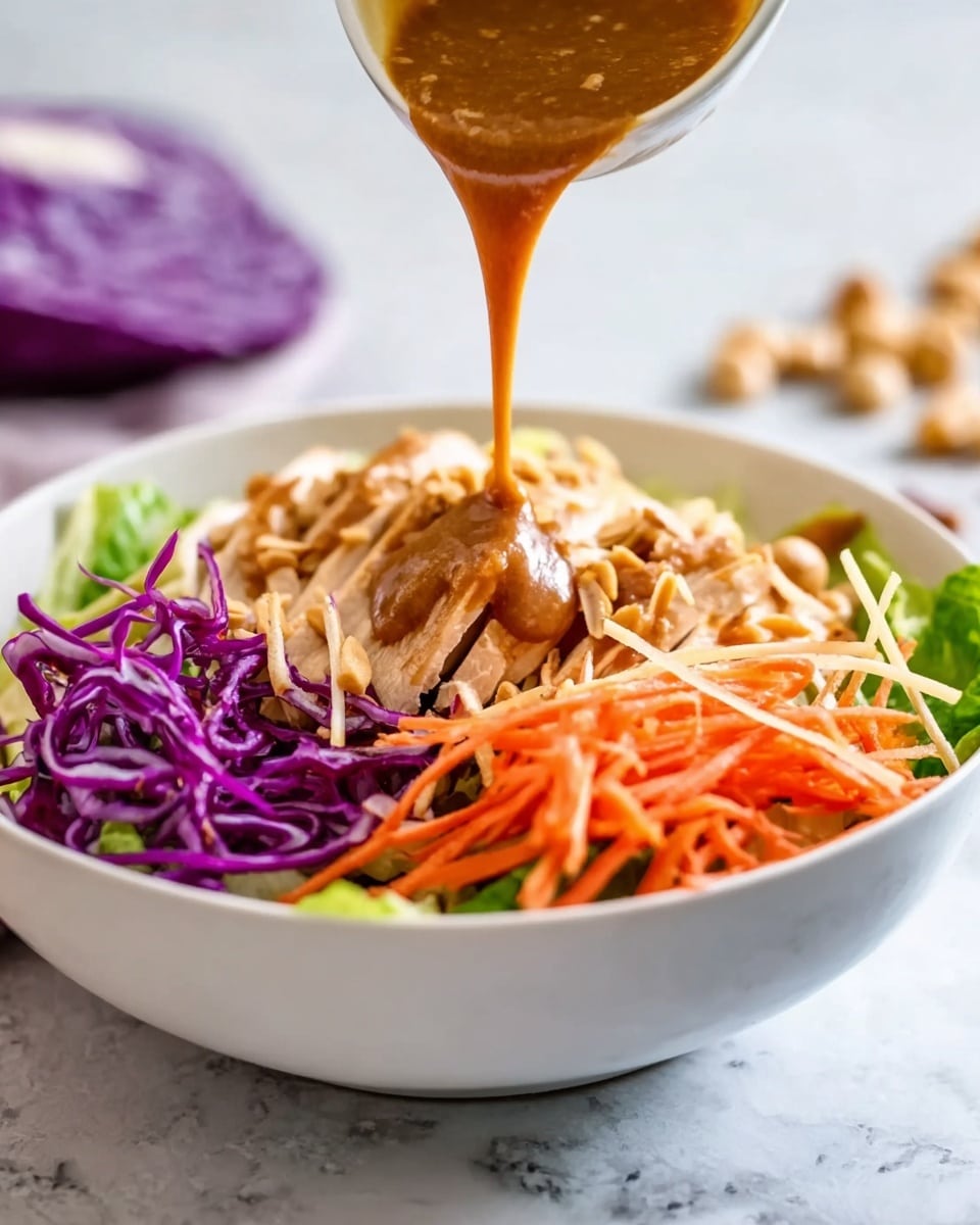 A white bowl filled with layers of food on a white marbled surface, starting with green lettuce at the bottom, topped with sliced cooked chicken pieces, thin orange carrot strips, and shredded purple cabbage on one side. A woman's hand is pouring thick brown sauce smoothly over the top layers, adding a glossy texture. Some small crunchy strips are sprinkled on top of the chicken. There is a blurred purple cabbage and some nuts in the background. Photo taken with an iphone --ar 4:5 --v 7