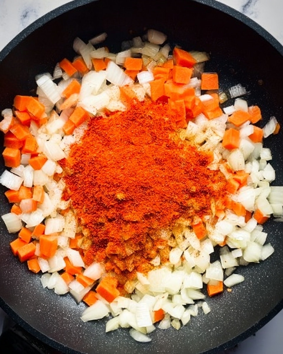 A close-up view of a black frying pan filled with diced white onions and small orange carrot pieces evenly spread across the bottom. In the center, a pile of bright red paprika powder sits on top of the vegetables, creating a vivid color contrast. The texture of the onions looks soft and slightly translucent, while the carrots maintain a firm, slightly glossy appearance. The pan surface has a slight shine, suggesting oil or moisture. The overall scene is set on a white marbled surface. photo taken with an iphone --ar 4:5 --v 7