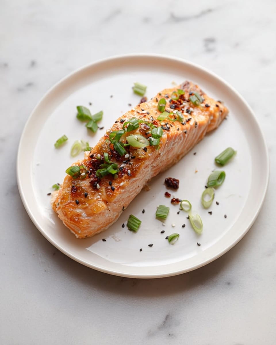 A single piece of cooked salmon with a golden-brown crust sits in the center of a round white plate, its flaky texture visible on the sides. The salmon is sprinkled with small pieces of green onion and black sesame seeds, adding bits of green and black color on top. Around the salmon, there are a few scattered herb leaves and small bits of chopped green onions on the plain white plate. The plate rests on a white marbled surface that adds a soft, elegant background to the dish. Photo taken with an iphone --ar 4:5 --v 7