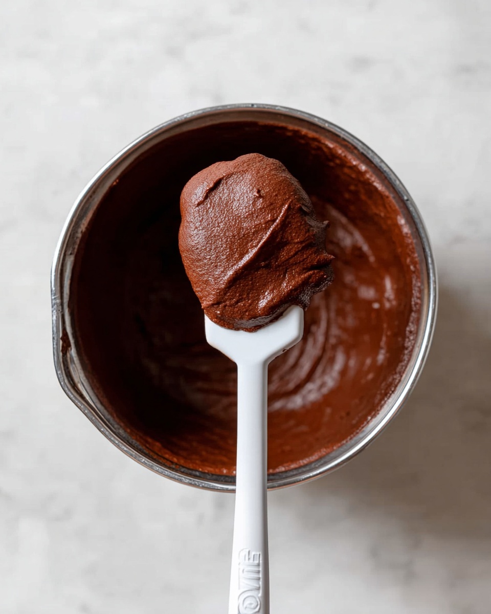20 Minute Fudgy Chocolate Brownie Cookies Recipe 4 A close-up view of a metal bowl filled with smooth, rich dark brown chocolate mixture. A white spatula rests on top of the bowl, holding a rounded scoop of the thick, creamy chocolate batter, showing its dense and slightly glossy texture. The scene is on a white marbled surface, giving a clean and bright background. photo taken with an iphone --ar 4:5 --v 7