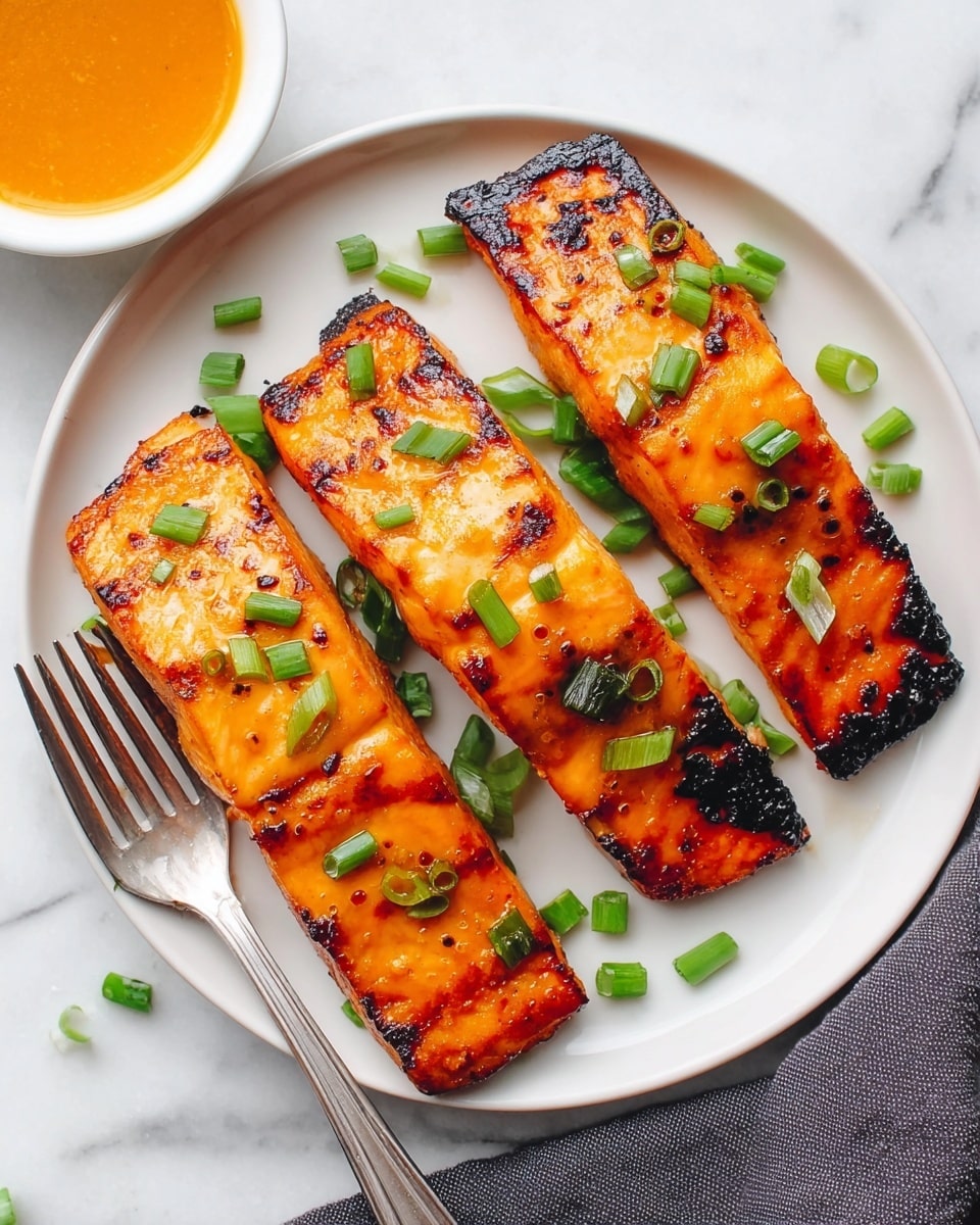 The image shows three pieces of grilled salmon with a shiny, orange glaze, each piece having slight char marks and a few darkened edges, arranged in a row on a white plate. Small green chopped scallions are sprinkled over the top and beside the salmon, adding a fresh green contrast. A silver fork is placed on the left side of the plate, resting partly under the first salmon piece. The plate sits on a white marbled surface with a dark cloth partially visible on the right side. In the upper left corner, there is a small white bowl filled with an orange sauce. Photo taken with an iphone --ar 4:5 --v 7