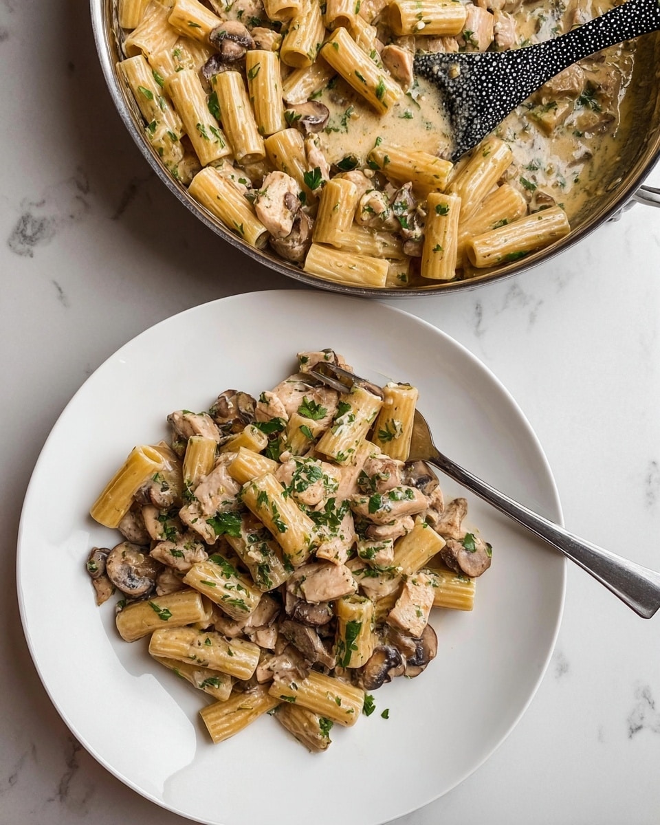 The image shows a white plate with rigatoni pasta served in a creamy sauce, mixed with sliced brown mushrooms and light brown cooked chicken pieces. The pasta and mushrooms are coated evenly with the creamy sauce and sprinkled with fresh green herbs on top. A silver fork rests on the right side of the pasta pile. Above the plate, there is a metal pan filled with the same rigatoni pasta, chicken, and mushrooms in the creamy sauce, along with a black and white patterned spoon resting inside. The background surface is a white marbled texture. Photo taken with an iphone --ar 4:5 --v 7