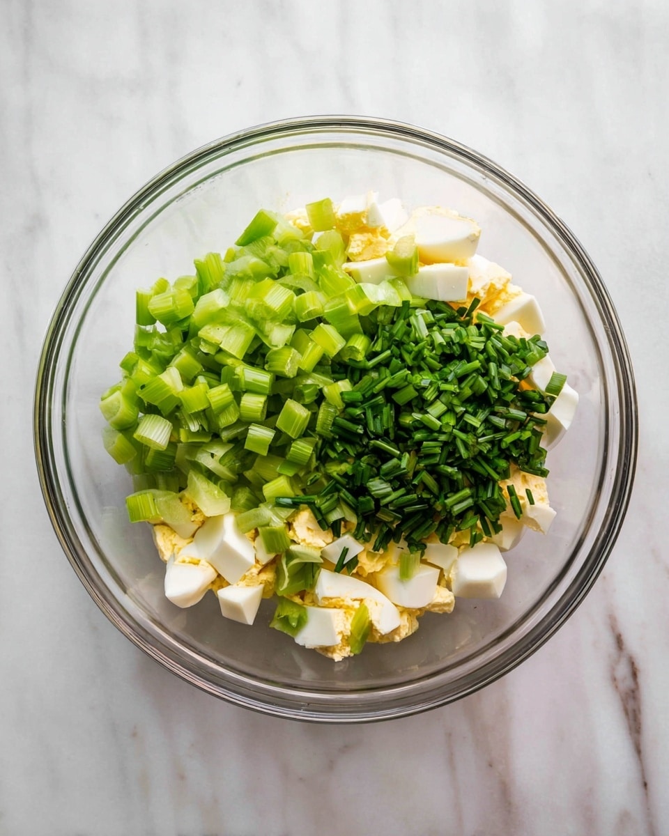 A clear glass bowl sits on a white marbled surface, holding three layers of chopped ingredients: at the bottom, there are pieces of white and pale yellow boiled eggs; on top of that, bright green celery pieces form a thick layer; and finally, a small pile of dark green chopped chives rests on one side of the bowl, providing a contrast of shapes and colors. photo taken with an iphone --ar 4:5 --v 7