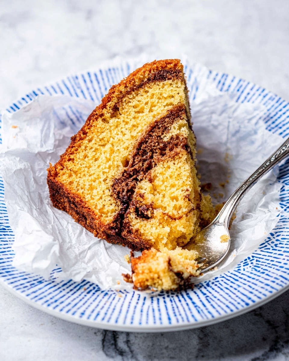A single slice of yellow cake with two dark brown swirls running through it, showing a soft crumb texture and a slightly darker, crispy crust around the edges. The slice sits on a crumpled piece of white parchment paper, placed on a white plate with a blue geometric pattern. A fork rests on the plate, with a piece of the cake on its tines. The whole scene is set on a white marbled surface. photo taken with an iphone --ar 4:5 --v 7