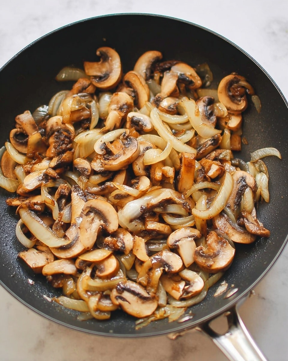 A black frying pan filled with cooked sliced mushrooms and onions. The mushrooms are light brown with some darker brown edges, showing a tender and slightly browned texture. The onions are translucent with a soft white and light golden color, mixed evenly among the mushrooms. The pan handle is visible on the right side, and the whole scene is set on a white marbled surface. Photo taken with an iphone --ar 4:5 --v 7