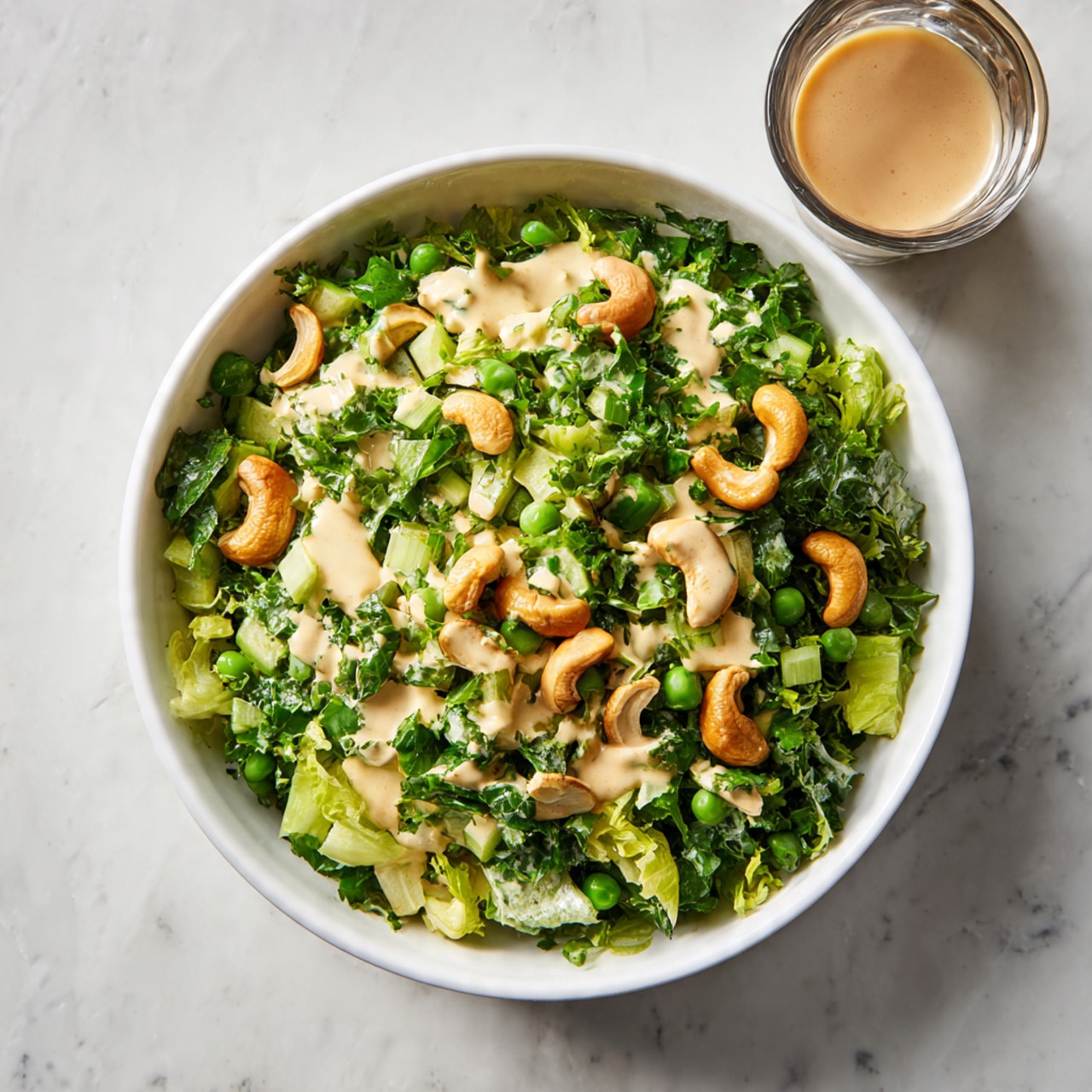The image shows a bowl filled with a fresh green salad. The base layer consists of small chopped green vegetables, including peas and leafy greens, creating a vibrant green mixture. On top, there are scattered pale cashew nuts adding texture and light brown color. A creamy light dressing is drizzled unevenly over the salad, giving it a shiny and moist look. The bowl is white and set on a white marbled surface, with a small glass of light brown liquid partly visible at the edge of the frame. photo taken with an iphone --ar 4:5 --v 7