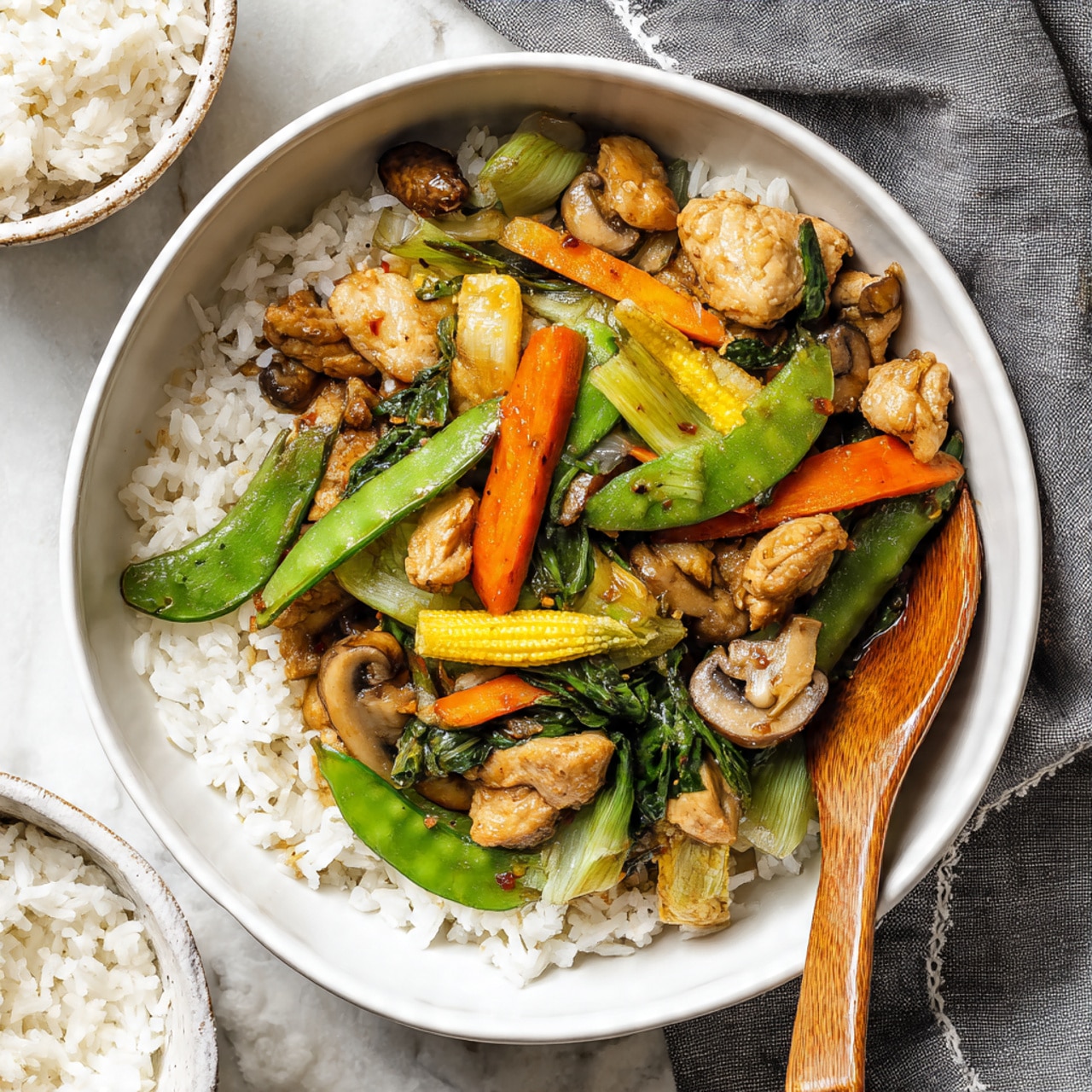 A white round bowl filled with a layer of white rice on the bottom, topped with a colorful stir-fry mix featuring light brown pieces of chicken, green sugar snap peas, yellow baby corn, orange carrot strips, green bok choy, and light brown mushroom slices, all coated lightly with sauce. A wooden spoon rests on the right side of the bowl, partially on the rice, with a white marbled textured surface underneath. In the upper left corner, a small round bowl holds more white rice. A gray cloth with white stitching is placed near the top right corner of the image. photo taken with an iphone --ar 4:5 --v 7
