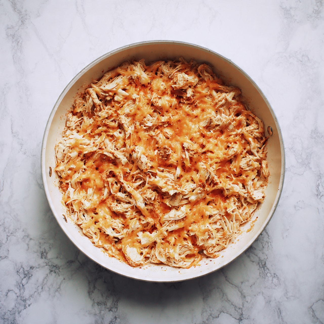 The image shows a white pan filled with shredded cooked chicken mixed with melted orange cheese evenly spread on top. The chicken appears tender and juicy, with the cheese slightly melted and blending into the meat. The pan is set on a white marbled surface, highlighting the warm colors of the chicken and cheese. The photo was taken with an iphone --ar 4:5 --v 7