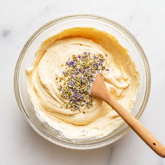 A clear glass bowl holds a creamy, light orange batter with a thick texture. In the center of the batter, there is a small pile of spices and finely chopped onions, adding colors of green, white, and purple. A wooden spatula with a light beige silicone head is resting inside the bowl, partially covered by the batter. The bowl is placed on a white marbled surface. photo taken with an iphone --ar 4:5 --v 7