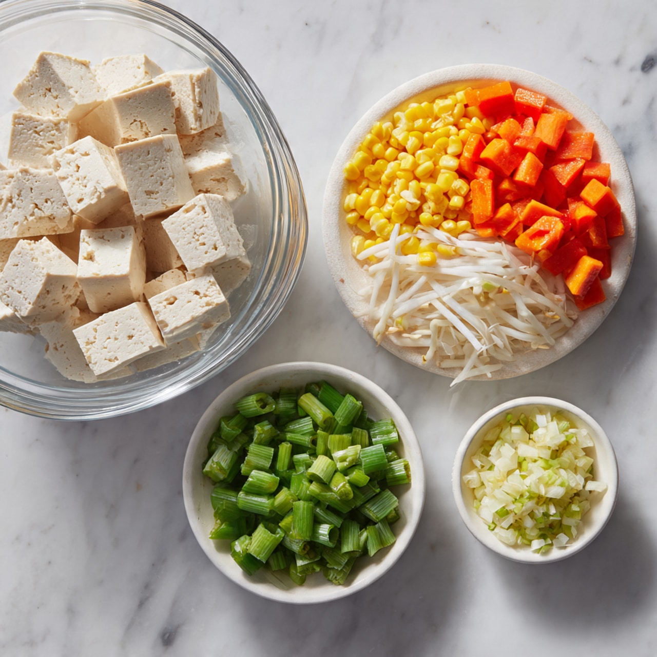 The image shows three white dishes on a white marbled surface. On the top left, a clear glass bowl contains large, crumbly chunks of pale beige tofu. Below the glass bowl, a round white dish holds six sections of chopped vegetables arranged separately: yellow corn kernels, orange diced carrots, white chopped onions, white bean sprouts, and bright green chopped long beans with small round pieces. On the right, a small white bowl has two piles of finely chopped ingredients; one is pale yellow minced garlic and the other is chopped green onions with white and green parts mixed. Photo taken with an iphone --ar 4:5 --v 7