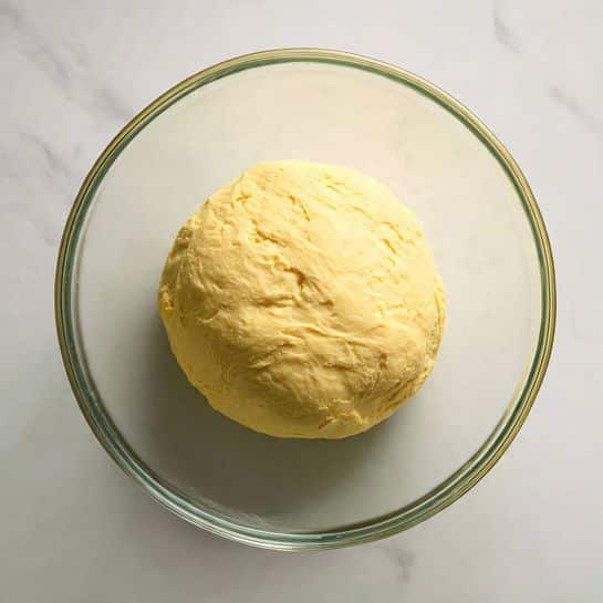 A single round ball of pale yellow dough with a slightly uneven surface sits in the middle of a clear glass bowl. The glass bowl is transparent and has smooth edges, placed on a clean white marbled surface. The dough looks soft with small folds and slight dimples on top, showing it has been rested or risen. There are no other objects or colors in the image, and the lighting makes the dough appear fresh and smooth. photo taken with an iphone --ar 4:5 --v 7