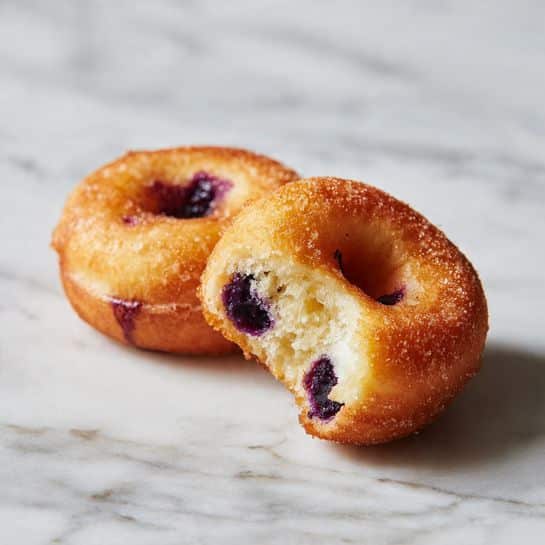 Two small donuts rest on a white marbled surface. They are light golden brown in color with a slightly rough texture, and both have small purple spots inside, suggesting blueberries. The donut in the front has a small bite taken out of it, showing the soft inside and more of the purple filling. The surface around the donuts looks smooth but has some natural marks. Photo taken with an iphone --ar 4:5 --v 7