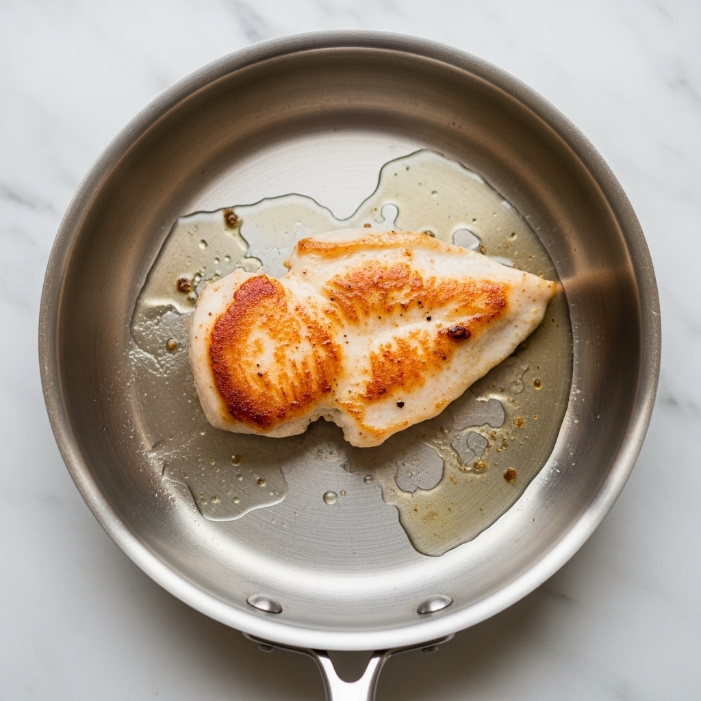 A single piece of lightly browned chicken is cooking in the center of a shiny stainless steel pan. The chicken has a golden, slightly uneven sear with some darker brown spots and is surrounded by a small pool of clear cooking oil. The pan is round with a simple handle extending out from the bottom. The whole pan is placed on a white marbled surface. photo taken with an iphone --ar 4:5 --v 7