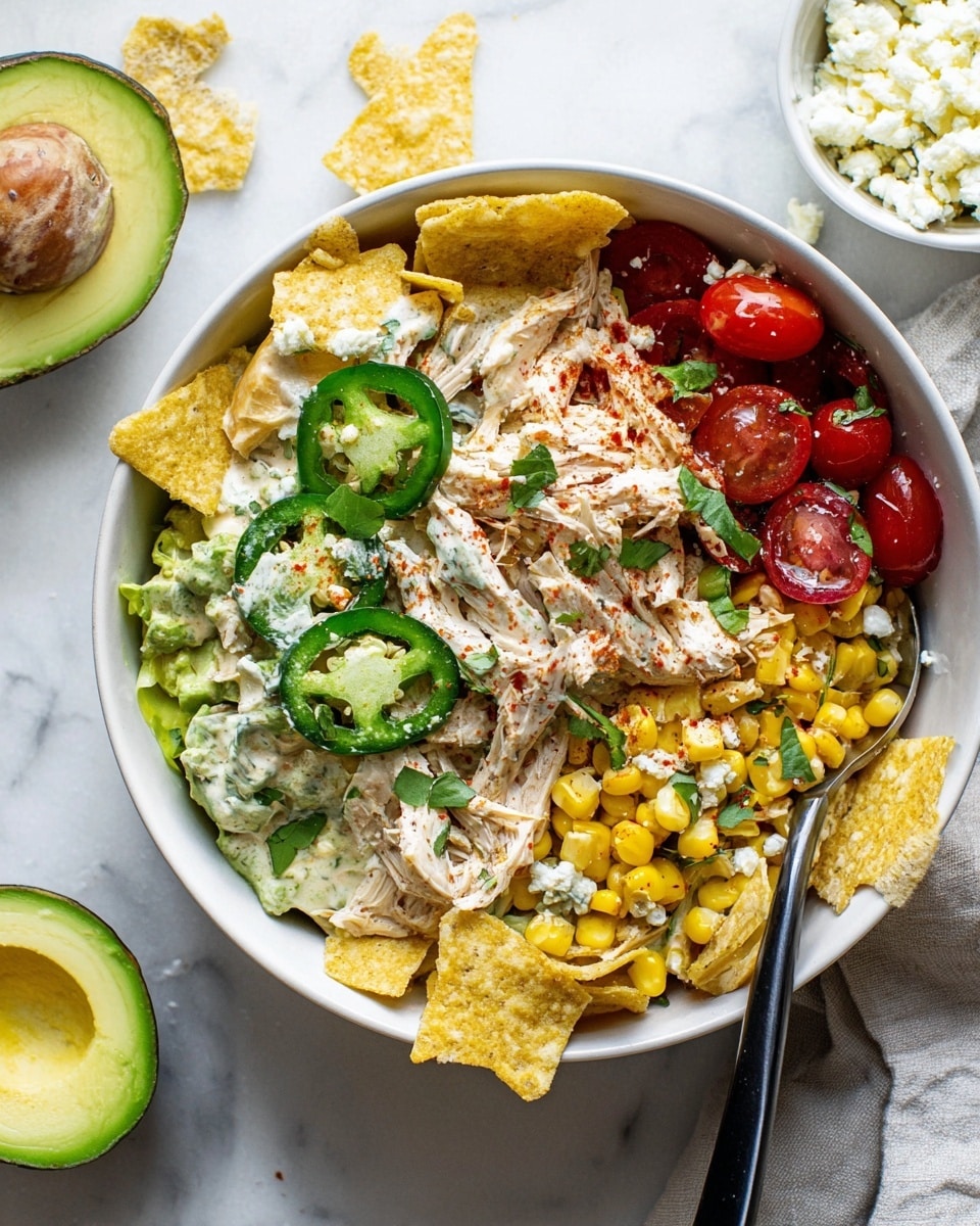 The image shows a white bowl filled with a layered salad on a white marbled surface. The bottom layer has crispy light yellow tortilla chips around the edges. Over the chips are sliced green avocado pieces dusted with red seasoning and scattered yellow corn kernels. Bright red cherry tomatoes add color in the middle. Large slices of white shredded chicken mixed with a creamy sauce form the main middle layer. On top, slices of green jalapeños with white crumbled cheese are placed beside more corn and small green herb leaves. A spoon with a black handle is standing inside the bowl near the right side. Nearby on the surface, there is a small white dish with white crumbled cheese and half of an avocado with a brown seed. Photo taken with an iphone --ar 4:5 --v 7