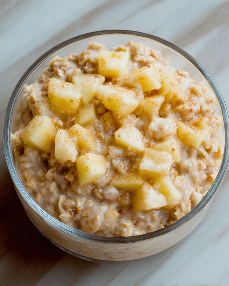 A close-up view of a clear glass bowl filled with creamy oatmeal mixed with small, soft chunks of light yellow apples. The oatmeal has a light brown color with a slightly thick and lumpy texture, showing some tiny specks of cinnamon or spice throughout. The bowl sits on a white marbled surface, making the oatmeal and apple pieces stand out clearly. The image captures the oatmeal from above in a simple and cozy setting, photo taken with an iphone --ar 4:5 --v 7