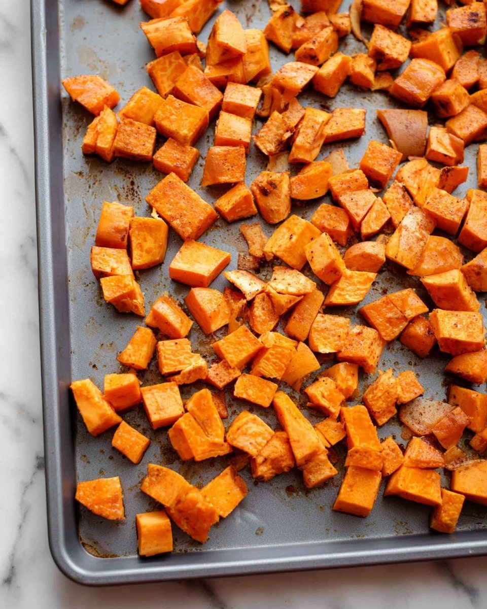 The image shows a gray baking tray filled with many pieces of roasted sweet potato. The pieces are cut into small, uneven cubes with soft edges and a bright orange color. Some pieces have light brown spots from baking, and a few are slightly darker or broken into smaller bits. Small drops of oil glisten on the sweet potatoes and the tray surface. The whole tray is placed on a white marbled surface. Photo taken with an iphone --ar 4:5 --v 7