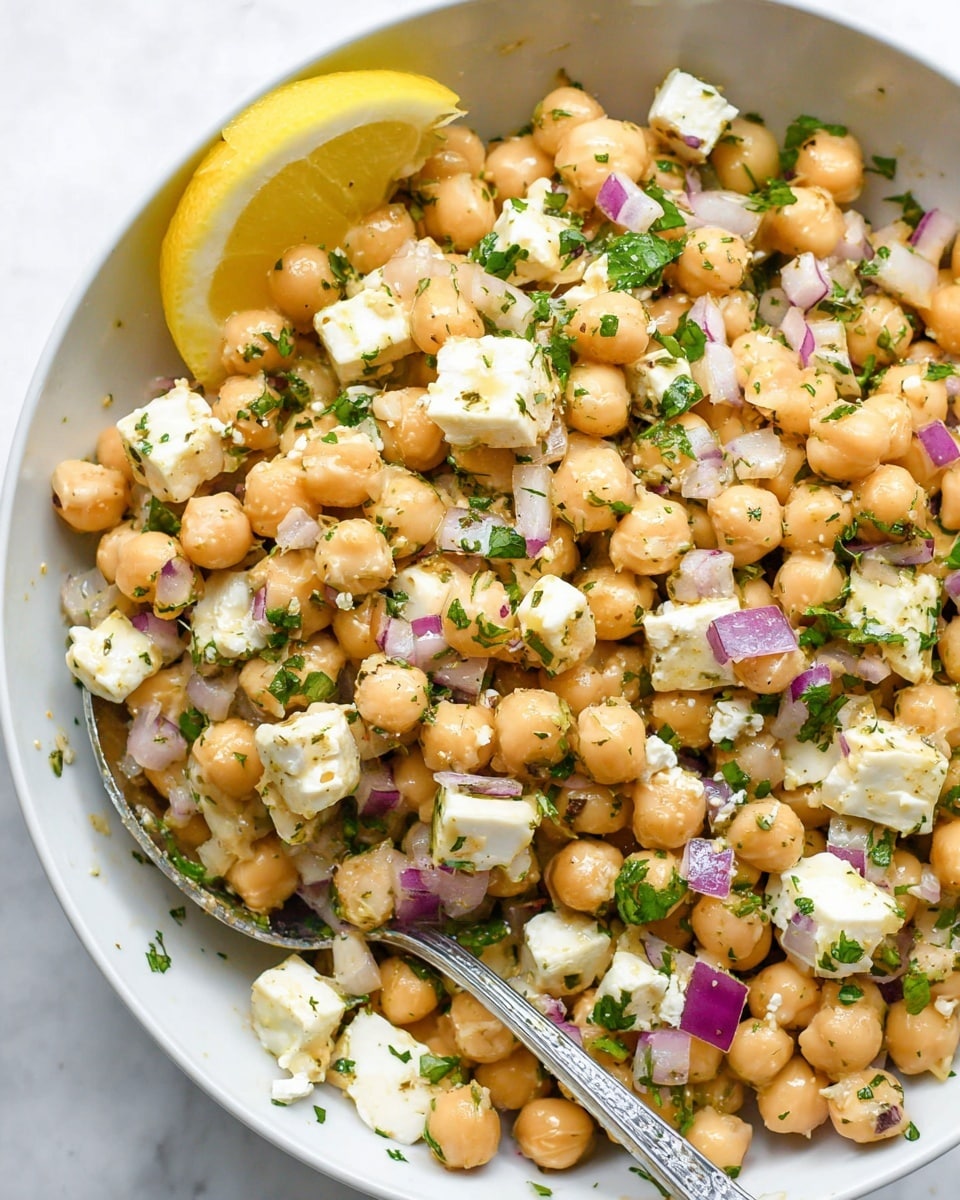 The image shows a close-up of a white bowl filled with a chickpea salad. The salad has three main layers: round beige chickpeas, small white cubes of cheese, and bits of purple onion scattered evenly on top. There are small green herb pieces mixed throughout, adding a fresh touch, and a silver spoon is partially buried in the salad on the right side. A lemon wedge with a bright yellow color is placed on the top left edge of the bowl. The background and surface beneath the bowl have a clean white marbled texture. photo taken with an iphone --ar 4:5 --v 7