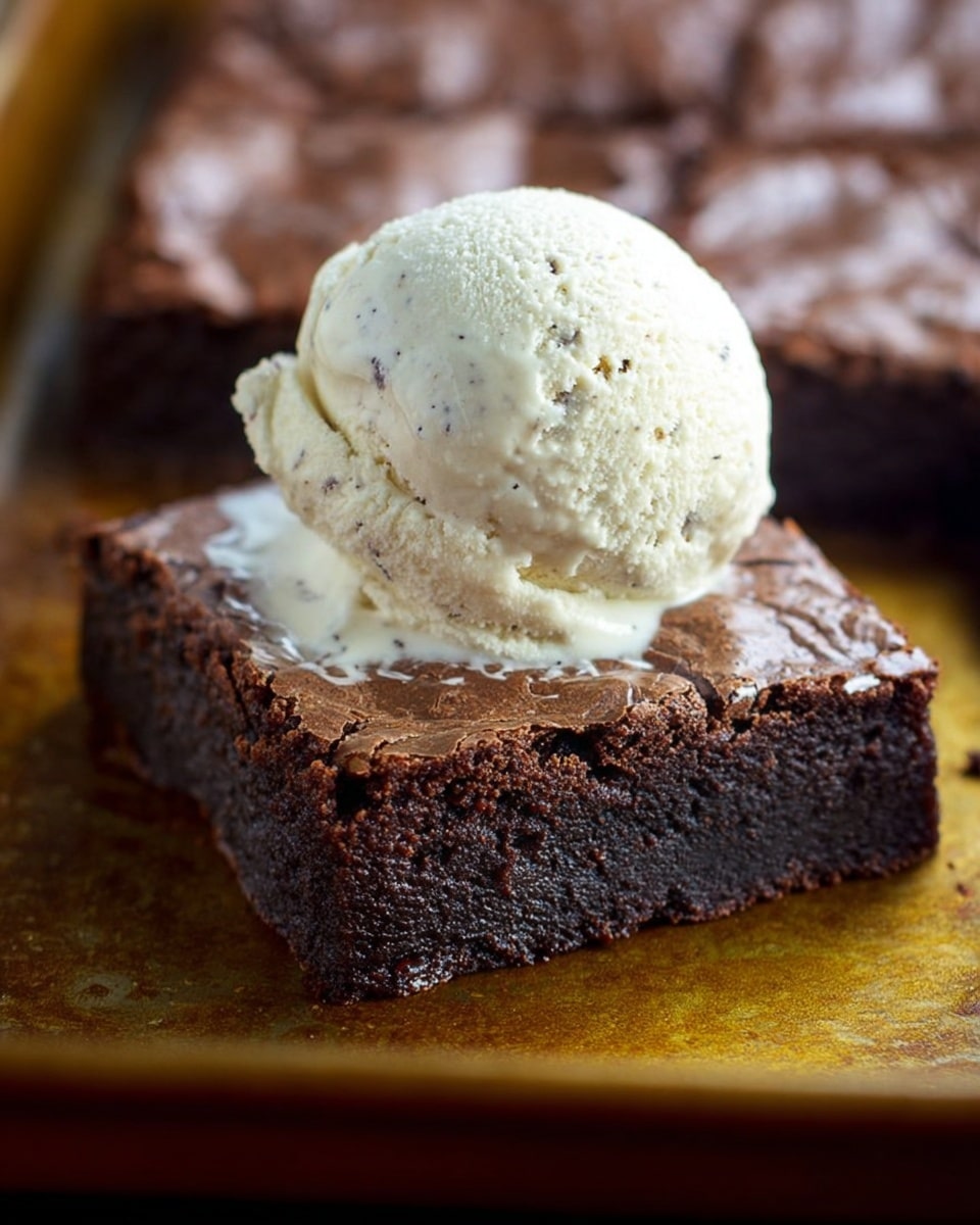 A close-up image of a square piece of dark brown chocolate brownie with a shiny, slightly cracked top layer. Sitting on top of the brownie is a single scoop of creamy off-white vanilla ice cream, showing tiny black vanilla specks and a smooth texture with some uneven edges. The brownie and ice cream rest on a baking tray with a warm golden surface, while other pieces of brownies are blurred in the background. photo taken with an iphone --ar 4:5 --v 7