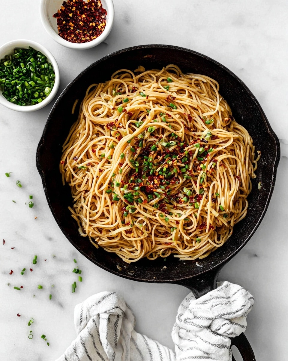 In a black cast iron pan, there is one layer of light brown noodles mixed with small red chili flakes and bright green chopped chives sprinkled evenly on top. The pan handle is wrapped in a white cloth with thin black stripes. The pan sits on a white marbled surface with two small white bowls near the top left, one containing more chopped green chives and the other filled with red chili flakes. Some scattered chives also lie on the surface around the pan. Photo taken with an iphone --ar 4:5 --v 7