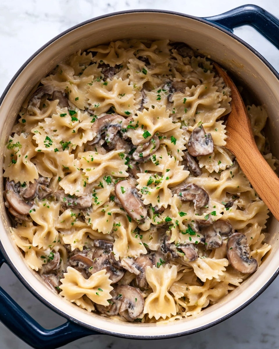 Inside a large white pot with a dark blue rim, there is a creamy pasta dish made of light beige bowtie pasta mixed with sliced brown mushrooms. The creamy sauce is smooth and coats the pasta evenly, giving it a shiny look. Small green herb pieces are scattered over the dish, adding a touch of color. On the right side, a wooden spoon is slightly submerged in the pasta, resting near the edge of the pot. The pot sits on a white marbled surface. photo taken with an iphone --ar 4:5 --v 7