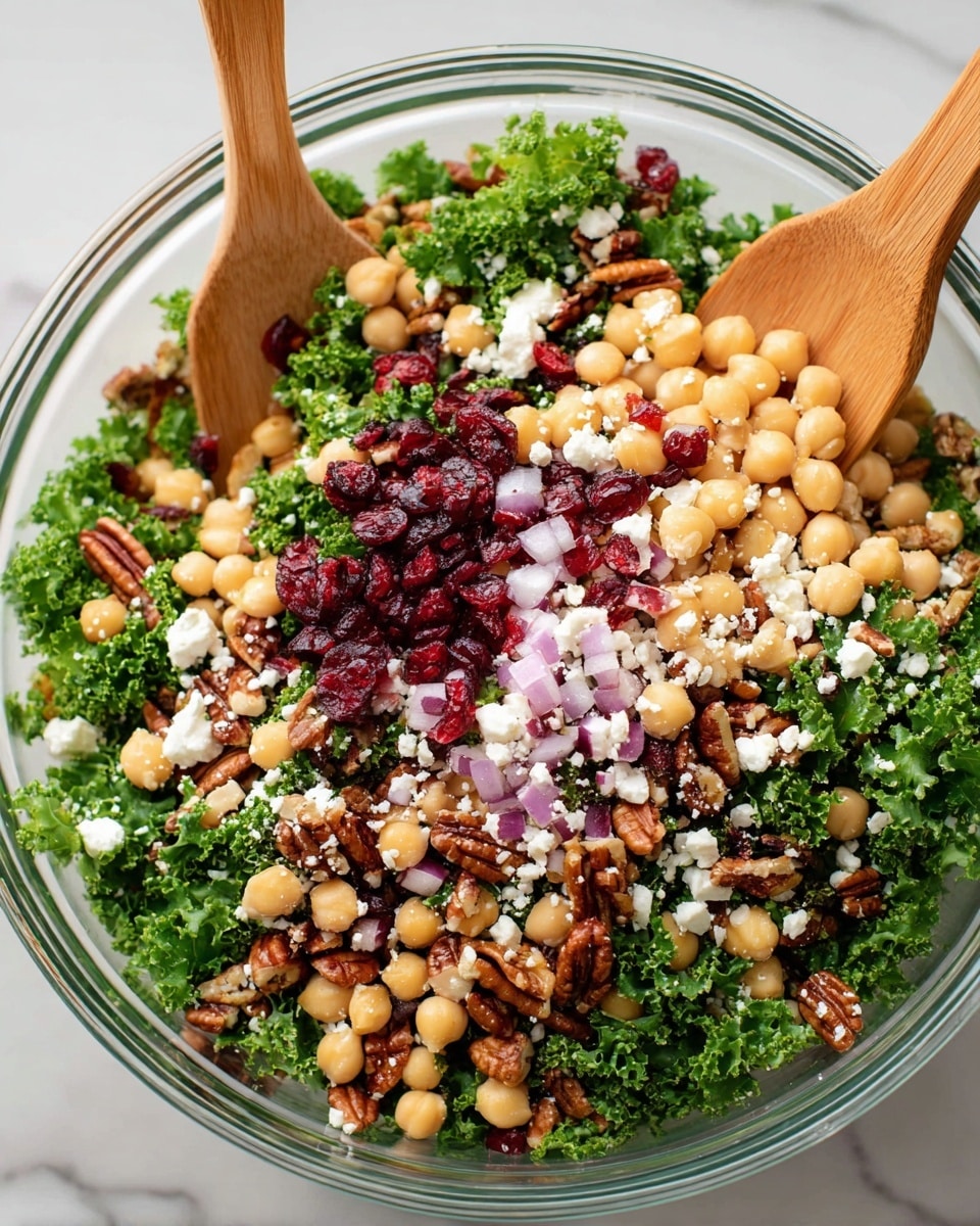 The image shows a clear glass bowl filled with a colorful salad placed on a white marbled surface. The salad has several layers: a bottom layer of green curly kale leaves, a middle layer of pale yellow chickpeas scattered throughout, and a layer of small red dried cranberries adding vibrant pops of color. There are also small pieces of chopped purple-red onions spread evenly on top, along with crumbled white feta cheese. Mixed in are medium-brown pecan halves, adding texture and contrast. Two wooden salad spoons are partially buried in the salad, one on the left and one on the right side of the bowl. Photo taken with an iphone --ar 4:5 --v 7