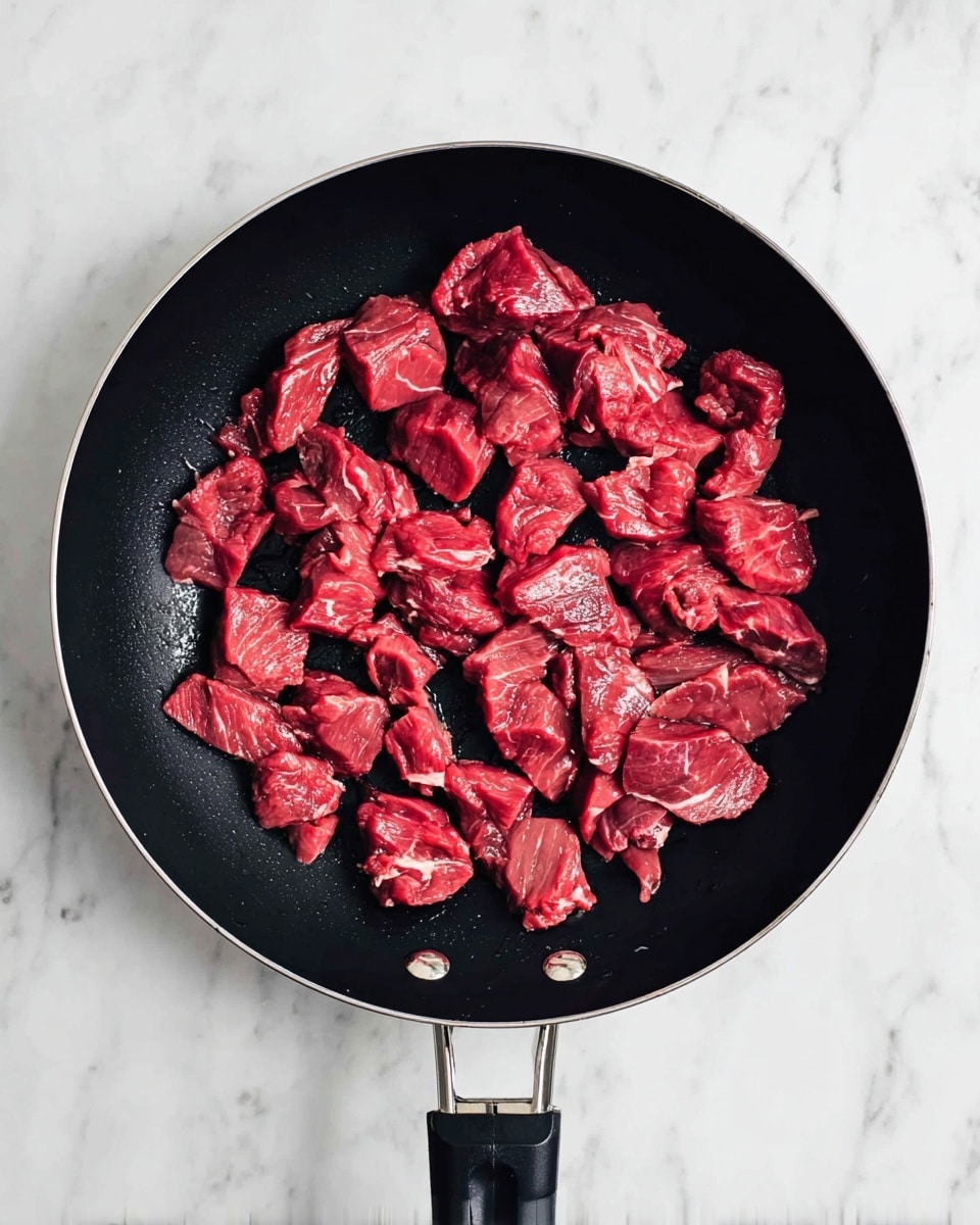 A black frying pan with a black handle is filled with small pieces of raw red meat spread out evenly inside the pan. The meat pieces vary in size and shape, showing smooth and slightly textured surfaces with some white fat spots. The pan rests on a white marbled surface, creating a clean and bright background. Photo taken with an iphone --ar 4:5 --v 7