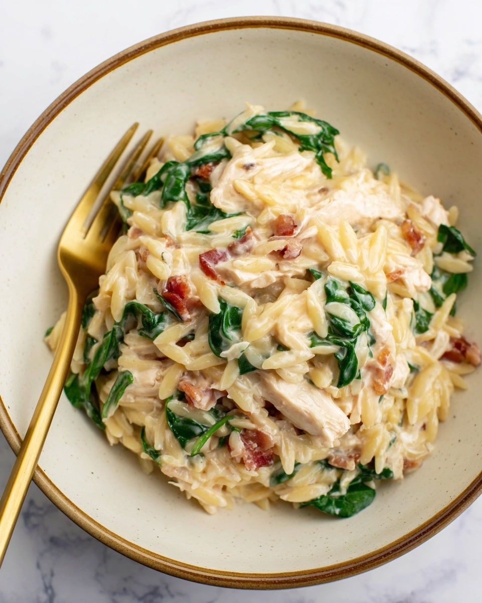 The dish shows one layer of creamy light yellow orzo pasta mixed with pieces of white shredded chicken, dark green spinach leaves, and small bits of light brown bacon. The ingredients are all mixed together, coated with a smooth white creamy sauce that makes the dish look rich and soft. The food is served in a round white bowl with a thin brown edge, placed on a white marbled surface. A gold and white fork is on the left side inside the bowl, adding a touch of elegance to the presentation. Photo taken with an iphone --ar 4:5 --v 7